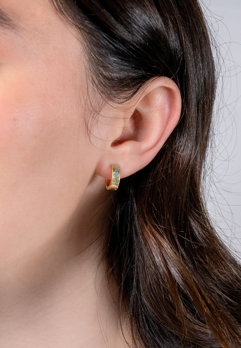 Goldene Creolen mit drei blauen Edelsteinen. Glatte Oberfläche und runde Form, die vom Ohrläppchen hängt. Strukturierte Details im Metall.