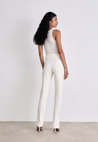 Weiße maßgeschneiderte Hose mit hoher Taille, kombiniert mit einem hellgrauen ärmellosen Oberteil. Das Model steht seitlich und zeigt geschmeidige, figurbetonte Linien.