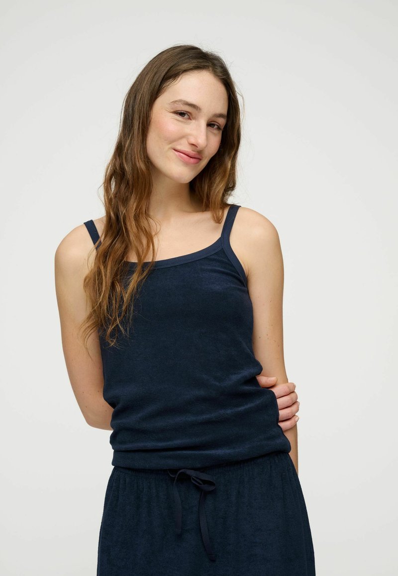 Femme aux longs cheveux ondulés portant un haut sans manches bleu marine et un pantalon à cordon assorti, souriante avec une main dans le dos.