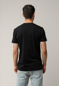 Schwarzes Baumwoll-T-Shirt mit kurzen Ärmeln, Rundhalsausschnitt und lässiger Passform, kombiniert mit hellblauen Jeans, die von der Taille abwärts sichtbar sind.