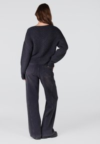 Dik, donkergrijs gebreid sweater met geribbelde textuur, gecombineerd met losvallende, donkergrijze denim broek en stevige zwarte schoenen.