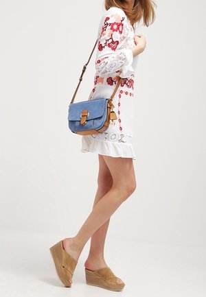 Blauwe suede schoudertas met een bruine leren band en sluiting; gedragen met een witte geborduurde jurk en tan kleurige instapschoenen.