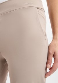 Pantalons beige taille haute en tissu lisse et extensible. Dotés d'une poche latérale et d'un logo en relief sur la ceinture.