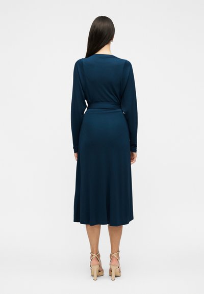 Γυναίκα με μακριά σκούρα μαλλιά, ντυμένη με μακρυμάνικο μίντι φόρεμα σκούρου μπλε χρώματος και μπεζ ψηλοτάκουνα σανδάλια, στέκεται μπροστά σε απλό φόντο.