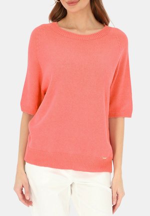 Kobieta ubrana w koralowy, krótki sweter z dzianiny z krótkim rękawem, zestawiony z białymi spodniami, na jednolitym tle.