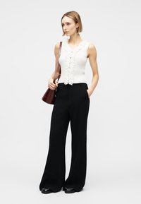 Γυναίκα όρθια, φοράει λευκή αμάνικη μπλούζα με βολάν, μαύρο παντελόνι φαρδύ, μαύρα παπούτσια και κρατάει μια σκούρα καφέ τσάντα ώμου.