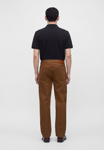 Homme portant une chemise noire à manches courtes, un pantalon marron à motifs et des chaussures foncées, debout, dos face à un fond clair uni.