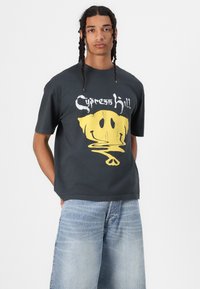 Čierne bavlnené tričko s veľkým grafickým motívom žltého úsmevu s efektem kvapkania a textom "Cypress Hill" nad ním. Voľný strih.