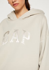 Hellbeige Kapuzenpullover aus weichem Stoff mit einer Kapuze und großem, reflektierendem "GAP"-Logo auf der Vorderseite. Einfaches, lässiges Design.