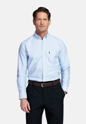 Man staat met handen gedeeltelijk in zakken, draagt een lichtblauw overhemd met knopen, ingebakken in zwarte broek met een bruine riem, neutrale achtergrond.