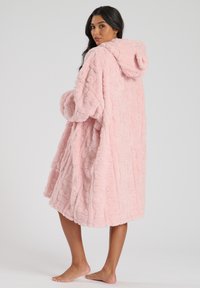 Coperta con cappuccio rosa, tessuto peluche testurizzato con un motivo ondulato, vestibilità oversize e maniche lunghe, che arriva a metà polpaccio, piedi nudi visibili.