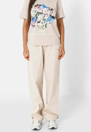 Beige katoenen t-shirt met bloemenprint en tekst, gecombineerd met ruime beige broeken en witte sneakers met grijze accenten.