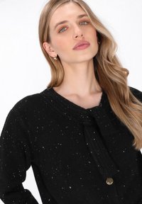 Zwarte gebreide cardigan met een gestructureerde afwerking, voorzien van een strik bij de kraag en een gouden knoopaccent. Gevlekte witte details doorheen.