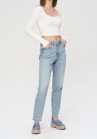 Witte ribgebreide langemouwen cropped top met knoopdetails, gecombineerd met lichtblauwe high-waisted jeans en blauwe platform loafers.