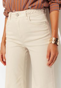 Pantalons taille haute de couleur crème avec un détail de ceinture tressée. Le modèle porte un bracelet métallique réfléchissant. Une chemise à rayures verticales est visible au-dessus.