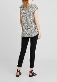 Donna che indossa una maglietta leopardata a maniche corte bianca, pantaloni skinny neri arrotolati e sandali neri con cinturini, in piedi contro uno sfondo bianco.