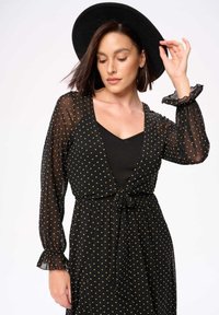 Zwart stippenjurk met doorschijnende lange mouwen, een diepe V-hals en een gestrikt middel. Accentueert met een brede zwarte hoed.