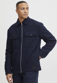 Jachetă din fleece bleu marin cu fermoar în faț�ă, două buzunare pe piept cu clapete și guler. Textură moale și croială ajustată.