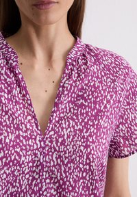 Lila Bluse mit V-Ausschnitt, kurzen Ärmeln und einem Muster aus weißen Sprenkeln. Weicher Stoff mit leicht gerafftem Detail am Ausschnitt.
