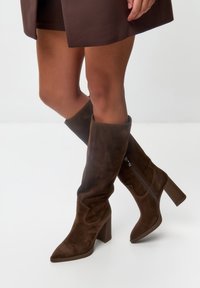 Stivali marroni al ginocchio realizzati in morbido suede, con punta affusolata, tacco a blocco e zip laterale per una facile vestibilità.