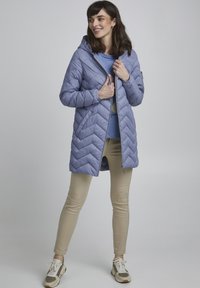 Cappotto imbottito viola chiaro con un motivo a zig-zag, cappuccio e chiusura con zip, indossato sopra una maglietta blu, abbinato a pantaloni beige e sneakers.
