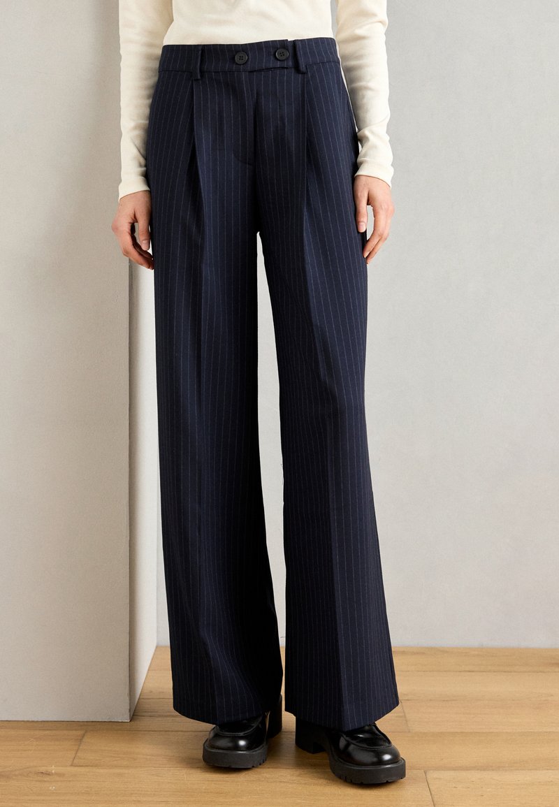 Pantalons rayés bleu marine à large jambe, dotés de deux boutons à la taille et d'une texture lisse et ajustée. Chaussures à plateforme noires.