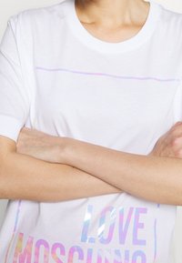 Bílé bavlněné tričko s krátkým rukávem, které má holografický nápis "LOVE MOSCHINO" a jemnou duhovou linku přes hrudník.