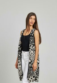 Schwarz- und cremfarbene Spitzenlongwesten mit aufwendigen floralen Mustern, offenem Design und ärmellosem Schnitt, getragen über einem schwarzen Top und weißen Hosen.