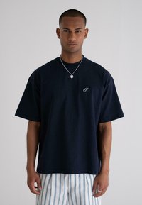 Navy blue cotton t-shirt with a relaxed fit, short sleeves, a crew neckline, and a small logo on the chest. Paired with striped pants.