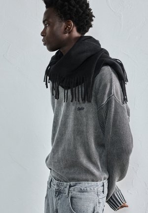 Jonge man met een donkere sjaal om zijn nek, gekleed in een gestructureerde grijze trui en lichtgrijze jeans, staand tegen een effen lichte muur.