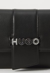 Juodas odinis rankinis krepšys, turintis stačiakampio formą. Apdailintas sidabriniu "HUGO" logotipu ir užlenkiamu uždarymu. Tekstūruotas paviršius su siūlų akcentais.