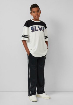 Weißes T-Shirt mit schwarzen Ärmeln und gestreiften Akzenten, mit buntem Text "SILVER." Kombiniert mit schwarzen Hosen und weißen Turnschuhen.