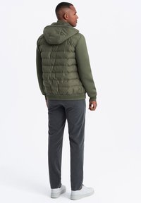 Olivgrön pufferväst med huva, med ett quiltat mönster, kombinerad med grå byxor. Vita sneakers fulländar outfiten.