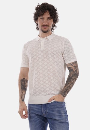 Uomo con capelli ricci e tatuaggi su entrambe le braccia, indossa una polo bianca a maniche corte con motivi e jeans blu, in piedi con una mano in tasca.