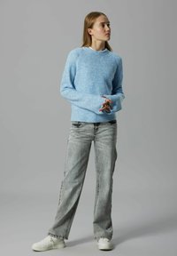 Młoda kobieta stoi z rękami złożonymi, ubrana w jasnoniebieski sweter, wyblakłe szare dżinsy i białe trampki na szarym tle.