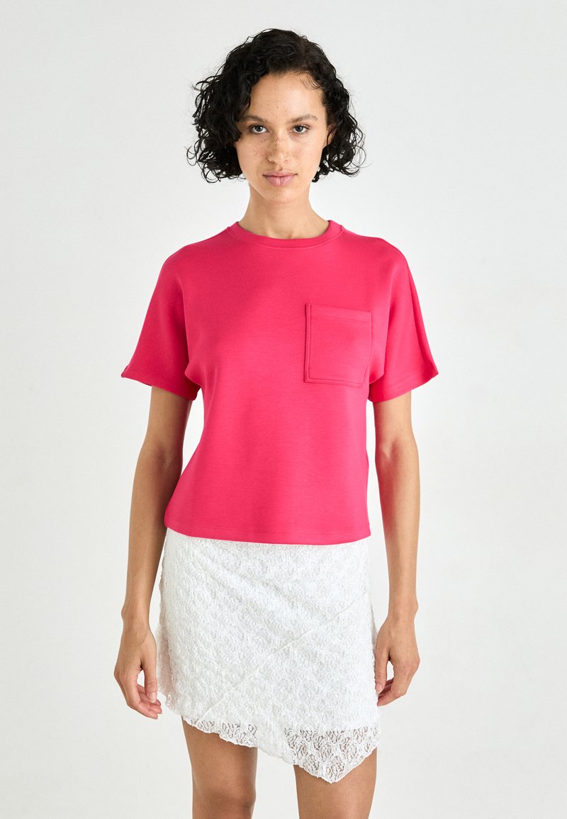 T-shirt en coton fuchsia à manches courtes avec une poche sur la poitrine, associé à une jupe en dentelle blanche de longueur genou avec un ourlet asymétrique.