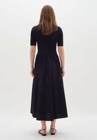 Schwarzes Kleid mit einem taillierten, gerippten Oberteil und einem fließenden, gerafften Rock. Kurzärmlig und knöchellang, kombiniert mit einfachen schwarzen Sandalen.