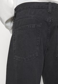 Jeans de denim gris oscuro con un diseño clásico, que presentan dos bolsillos traseros y una tela texturizada. Los detalles de la costura son visibles a lo largo de las costuras.