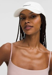 White baseball cap with "lululemon" logo, worn with a light pink tank top. Smooth texture and fitted design, showcasing a relaxed appearance.