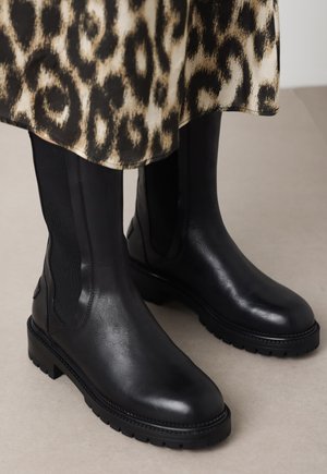 Zwarte leren enkellaarzen met elastische zijpanelen, stevige rubberen zolen en stiksels. Gepaard met een jurk met luipaardprint.