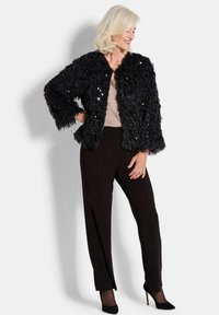 Schwarze, flauschige Jacke mit glänzenden Paillettenaussetzungen, kombiniert mit schwarzen Hosen und hohen Schuhen. Das Outfit hat ein strukturiertes, plüschiges Aussehen.