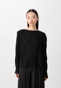 Top lungo nero a maniche lunghe con vestibilità morbida e scollatura a barca, caratterizzato da un tessuto liscio e delicate pieghe sulle spalle.
