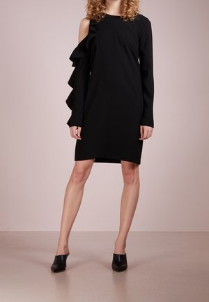 Frau trägt ein schwarzes knielanges Kleid mit einem langen Ärmel und Rüschendetail an der freiliegenden Schulter, kombiniert mit schwarzen mules mit Absatz.