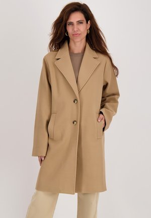 Vrouw met lang bruin haar, gekleed in een beige keurige jas tot op de knie met grote revers en twee knopen, met de handen in de zakken, staande tegen een effen achtergrond.
