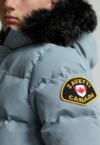 Veste doudoune bleu clair avec un col en fausse fourrure noire, arborant un logo circulaire avec une feuille d'érable et "Zavetti Canada" en jaune et noir.