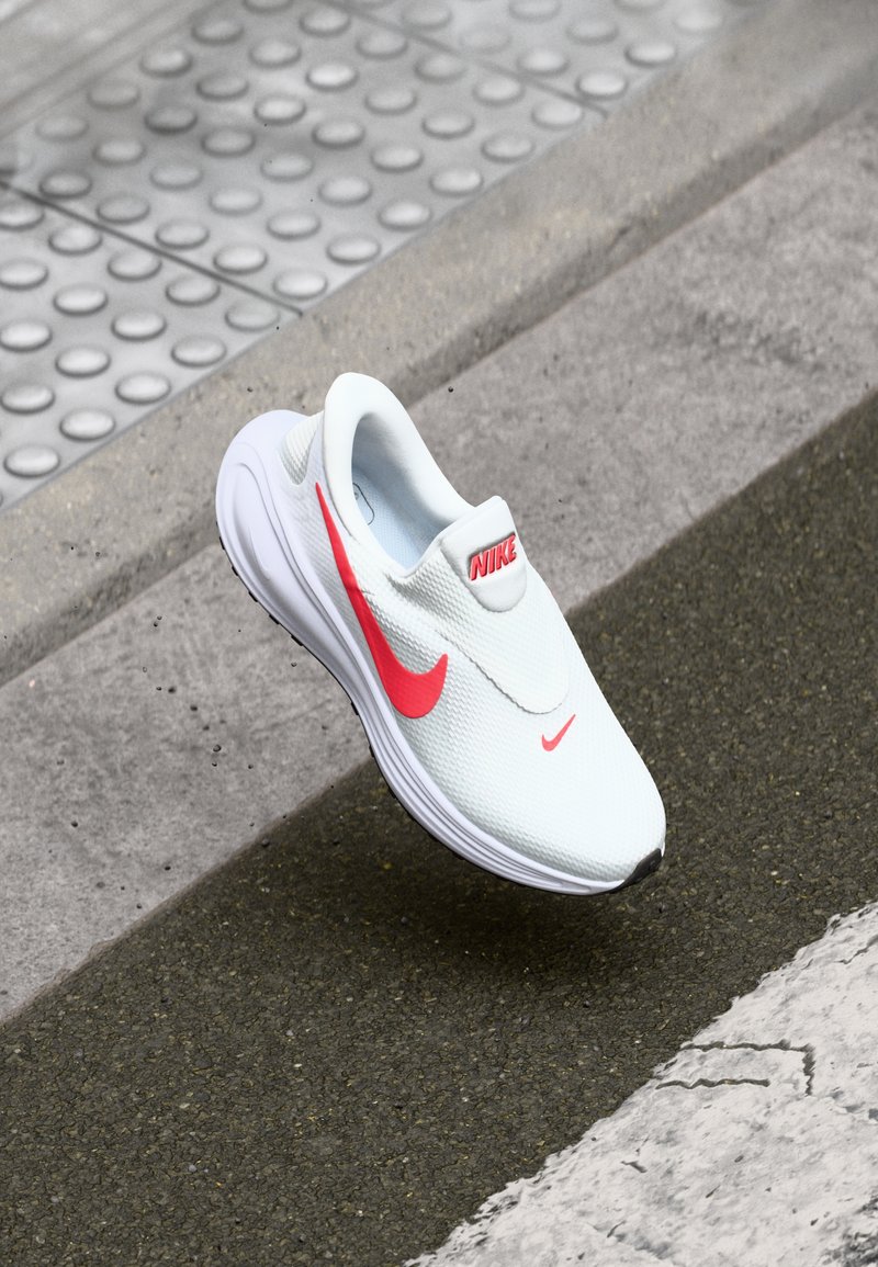 Λευκά slip-on sneakers της Nike με υφή πλέγματος, που διαθέτουν μια εμφανή ροζ σήμα και λογότυπο. Η σόλα είναι λεία με ελαφριά καμπυλότητα.