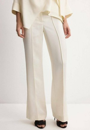 Mujer con pantalones anchos color crema y top a juego, de pie con zapatos de tacón abiertos negros, sobre un fondo liso.