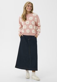 Világos rózsaszín pulóver fehér virágmintákkal, sötét farmer maxi szoknyával párosítva. Fehér sportcipők egészítik ki az öltözéket.