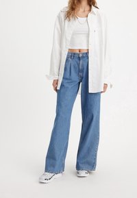 Vit långärmad skjorta med knappar över en vit crop top, tillsammans med ljusblå vida jeans och vita sneakers med svarta detaljer.