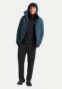 Giacca imbottita verde petrolio scuro con cappuccio, pile nero con mezza cerniera sotto, e pantaloni neri. La persona è in piedi con un'espressione neutra.